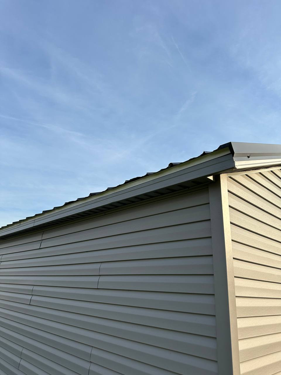 Corner of a light gray siding shed with horizontal panels and a metal roof edge against a clear blue sky