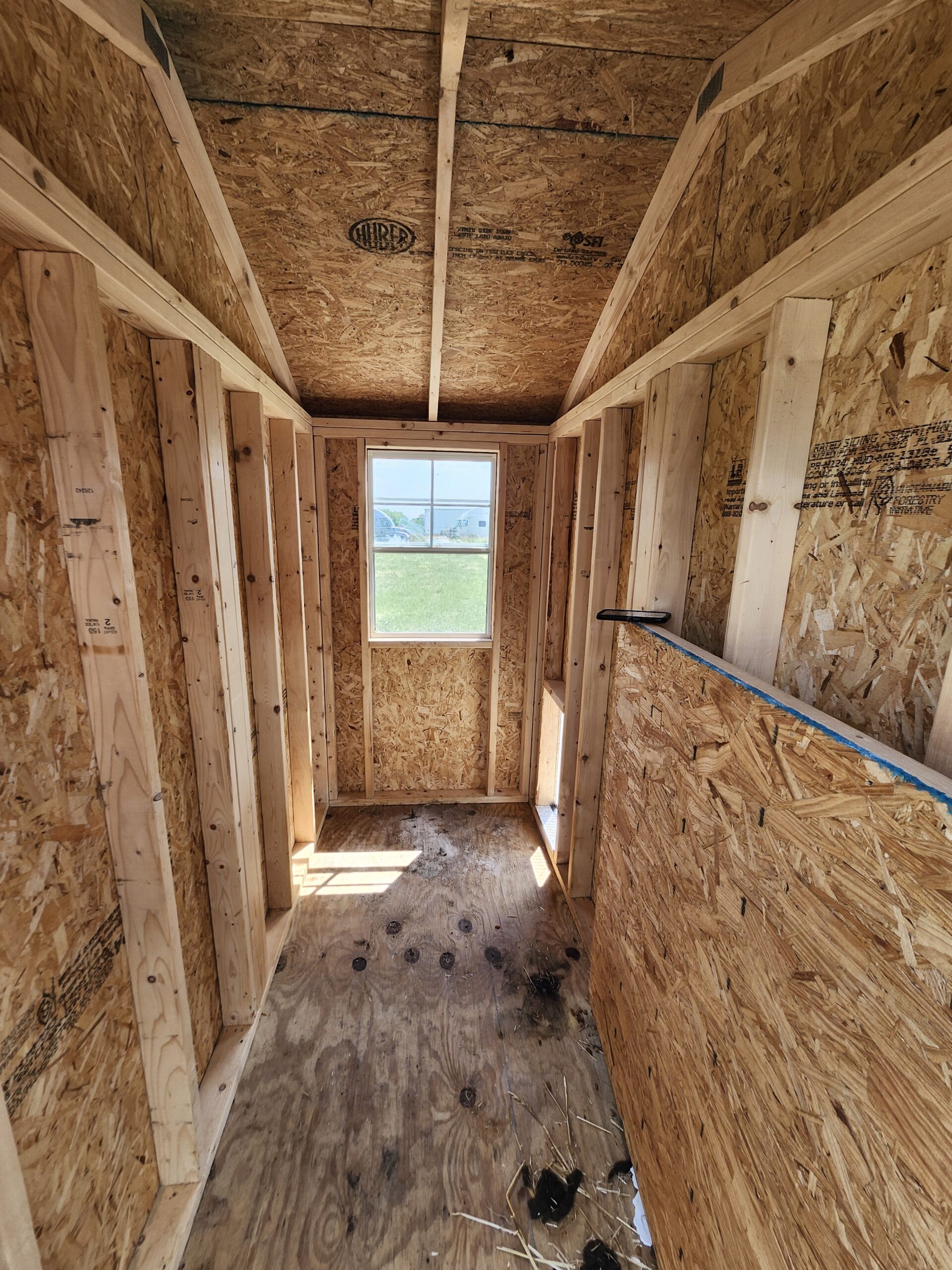 Interior of an unfinished, wood-framed room with OSB walls and exposed ceiling joists, a small window at the far end, and construction debris on the floor