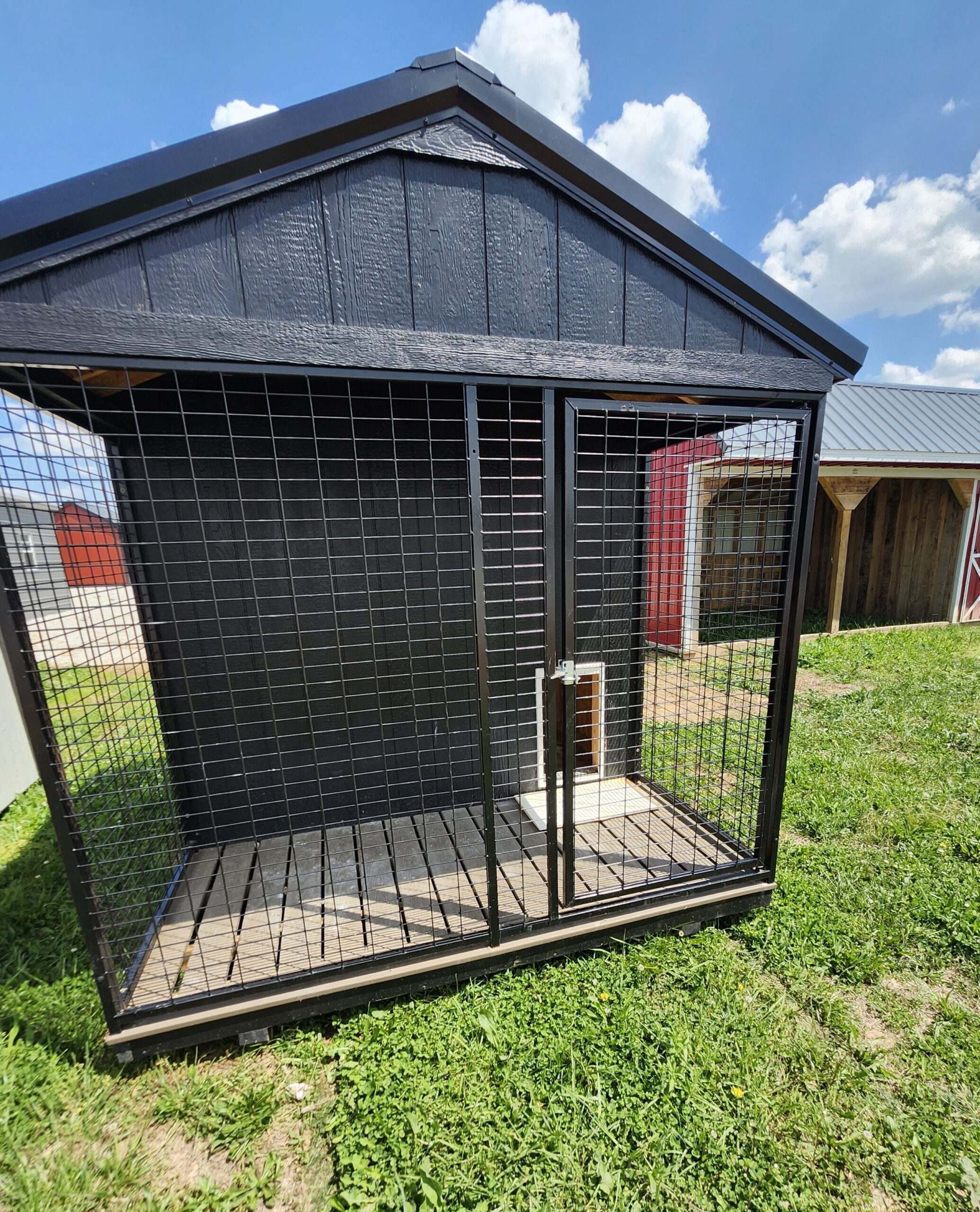 Black metal outdoor dog kennel with wire mesh panels standing on grass, a small doorway visible under a blue sky with clouds