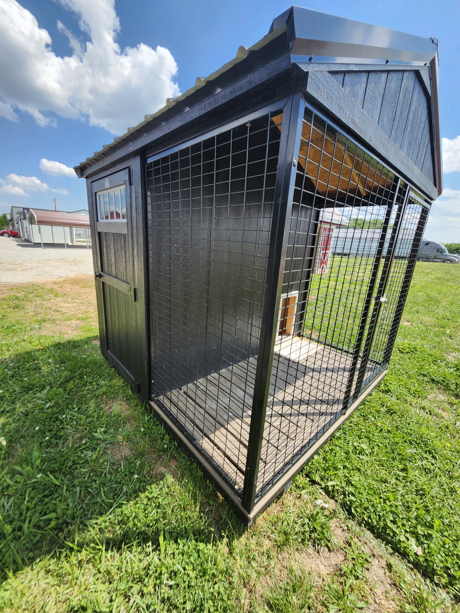 Outdoor black metal dog kennel with wire mesh sides on grassy field, clear blue sky in background.