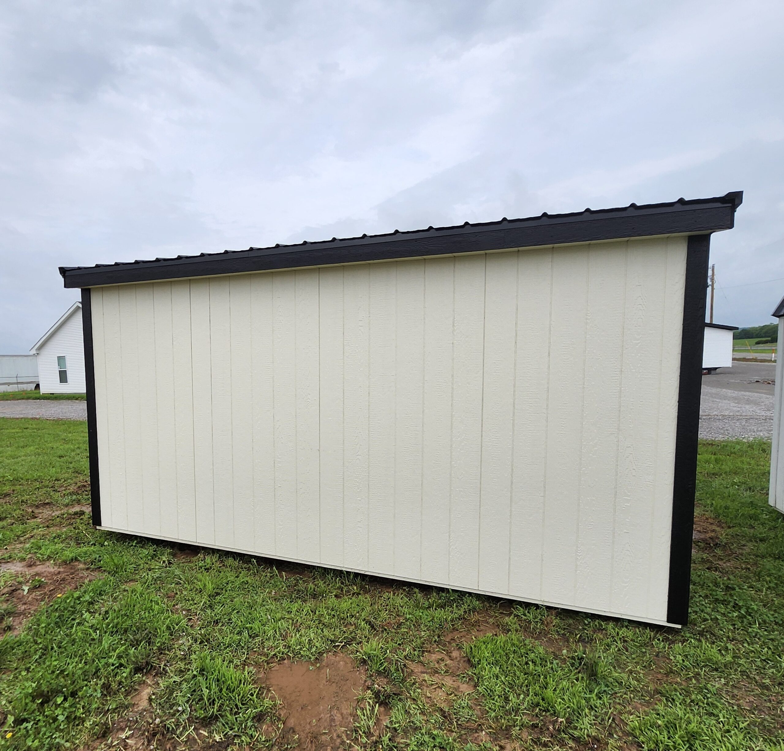 Beige shed with dark trim along the roofline in a grassy, outdoor area under a cloudy sky.