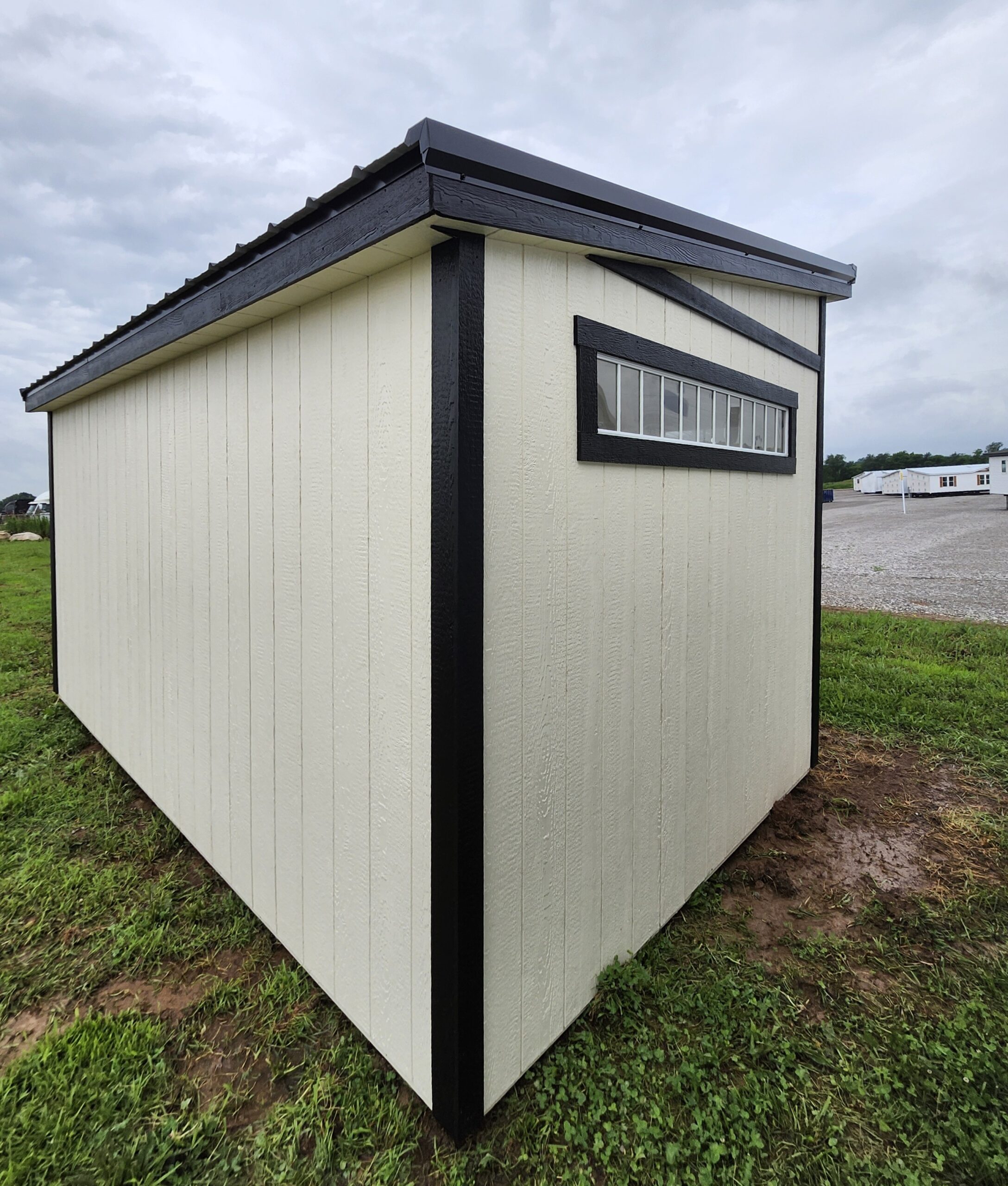 Beige storage shed with black trim and a long horizontal window, set on grass beside a gravel area under a cloudy sky.