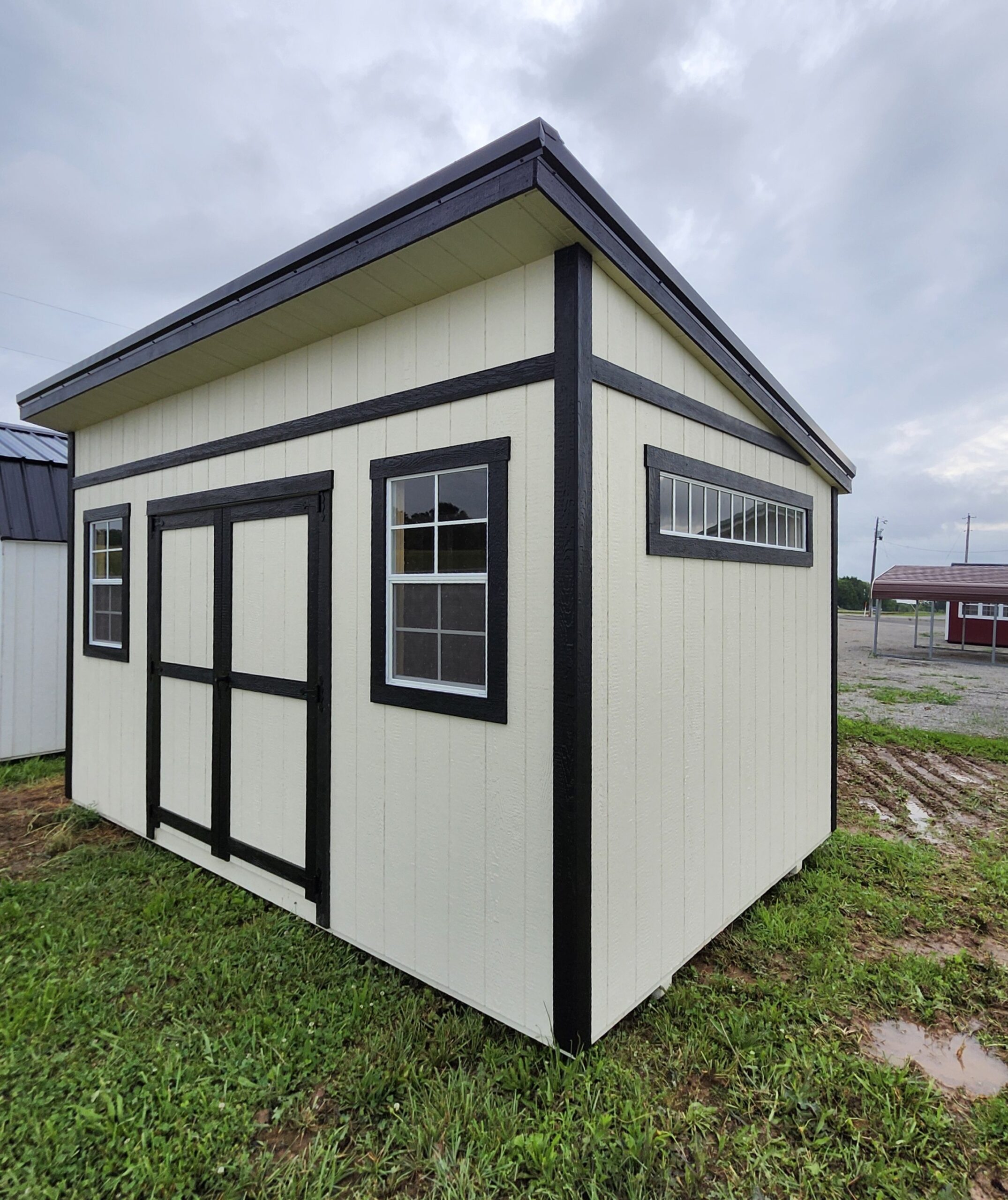 Small cream-colored shed with black trim and two windows, set on a grassy lot under a cloudy sky (corner view).