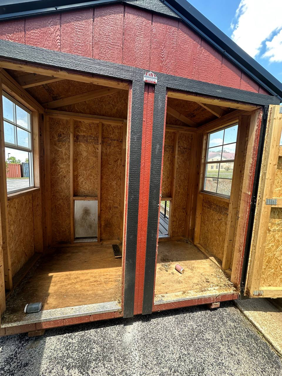 Open shed under construction with red siding and black trim, showing unfinished plywood interior and two windowed walls on either side.