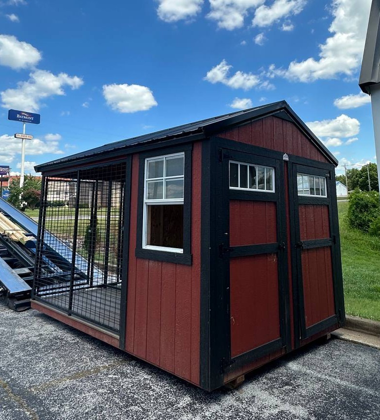 Red wooden chicken coop with black trim and a wire enclosed run, placed on a paved lot under a clear blue sky with fluffy clouds.