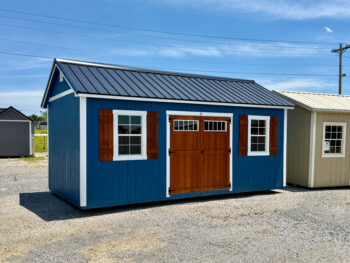 Blue shed with white trim, brown double doors, and a small window on a gravel lot under a clear blue sky.