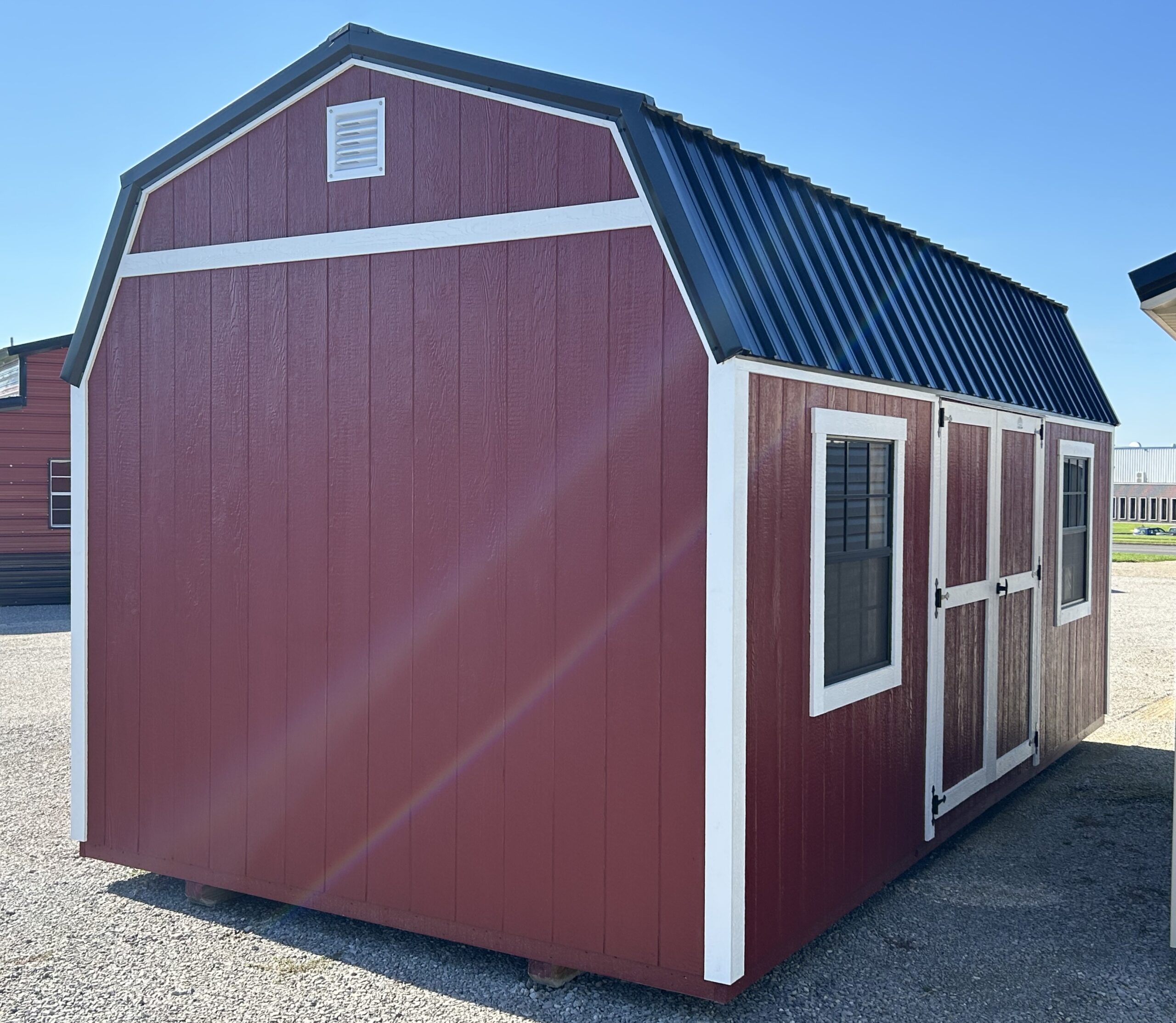 Red wooden shed with white trim and a black metal roof, set on a gravel lot under a clear blue sky.