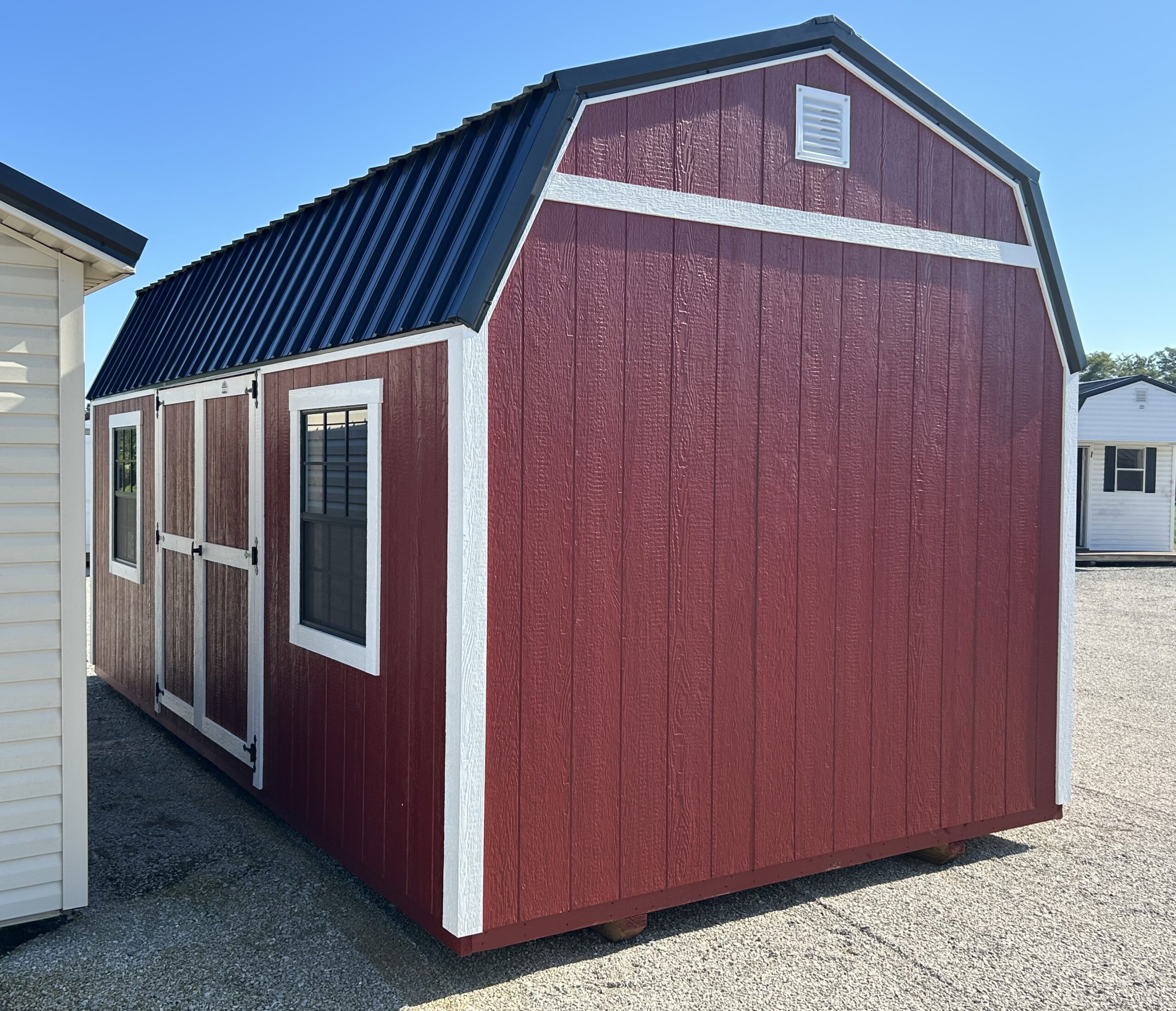 Red wooden storage shed with white trim, a gabled metal roof, and a small vent near the peak against a clear blue sky.