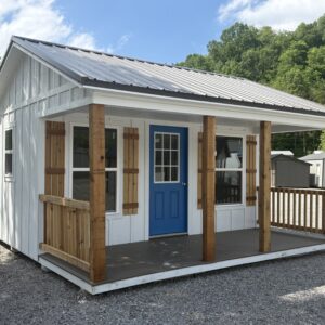 Small white wooden cottage with a bright blue door, covered porch, and wooden posts in a gravel lot with trees in the background.