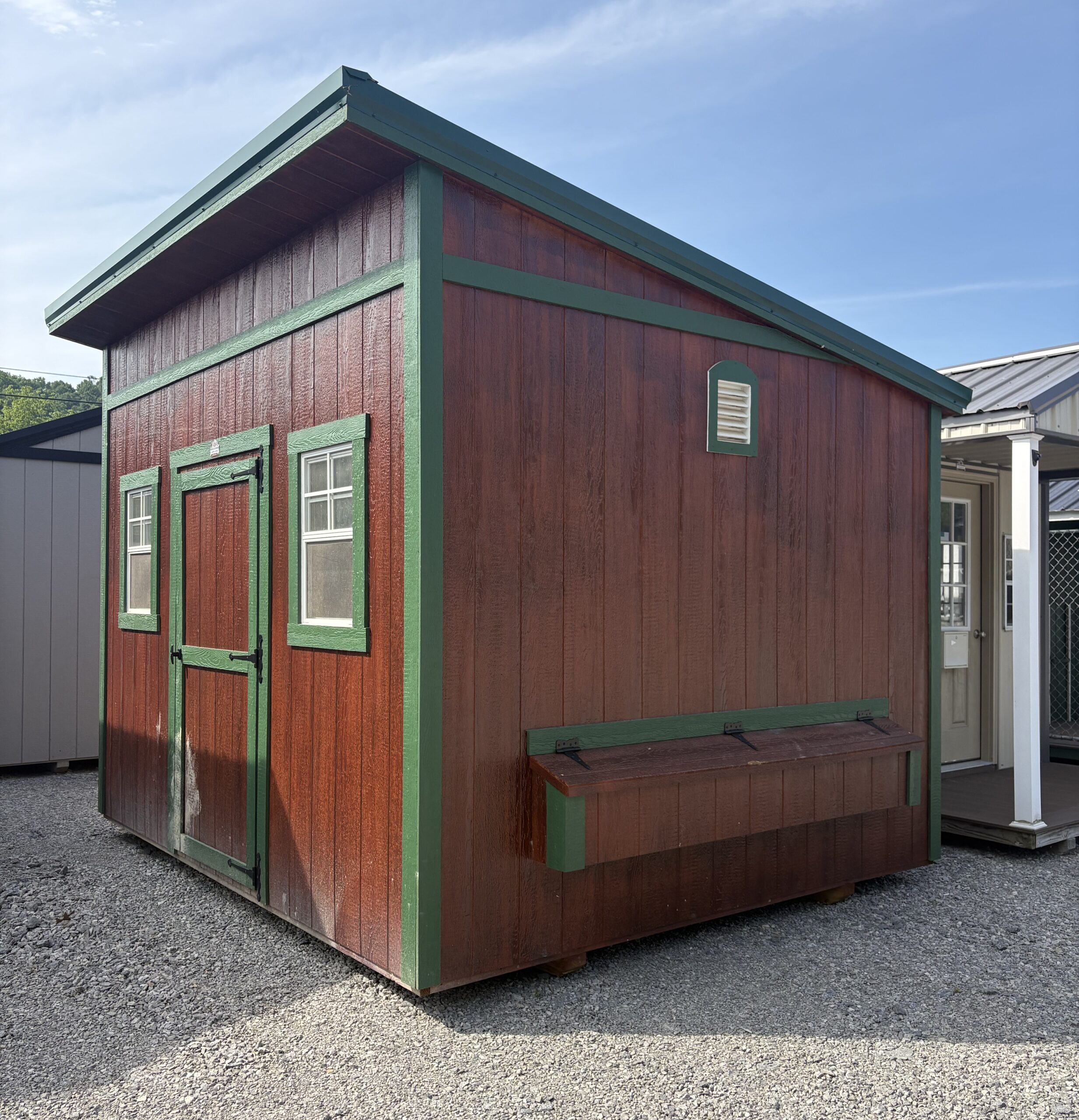 Wood storage shed with red walls and green trim on a gravel lot, showing two windows and a small overhanging roof.