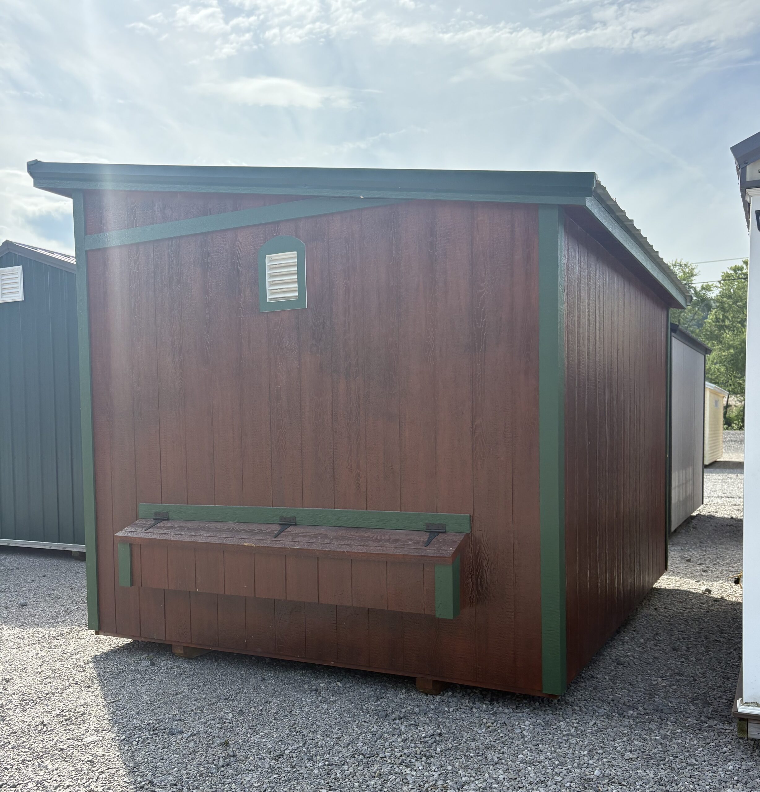 Brown wooden shed with teal-green trim, vent in wall, and a long wooden bench attached, outdoors on a gravel lot.
