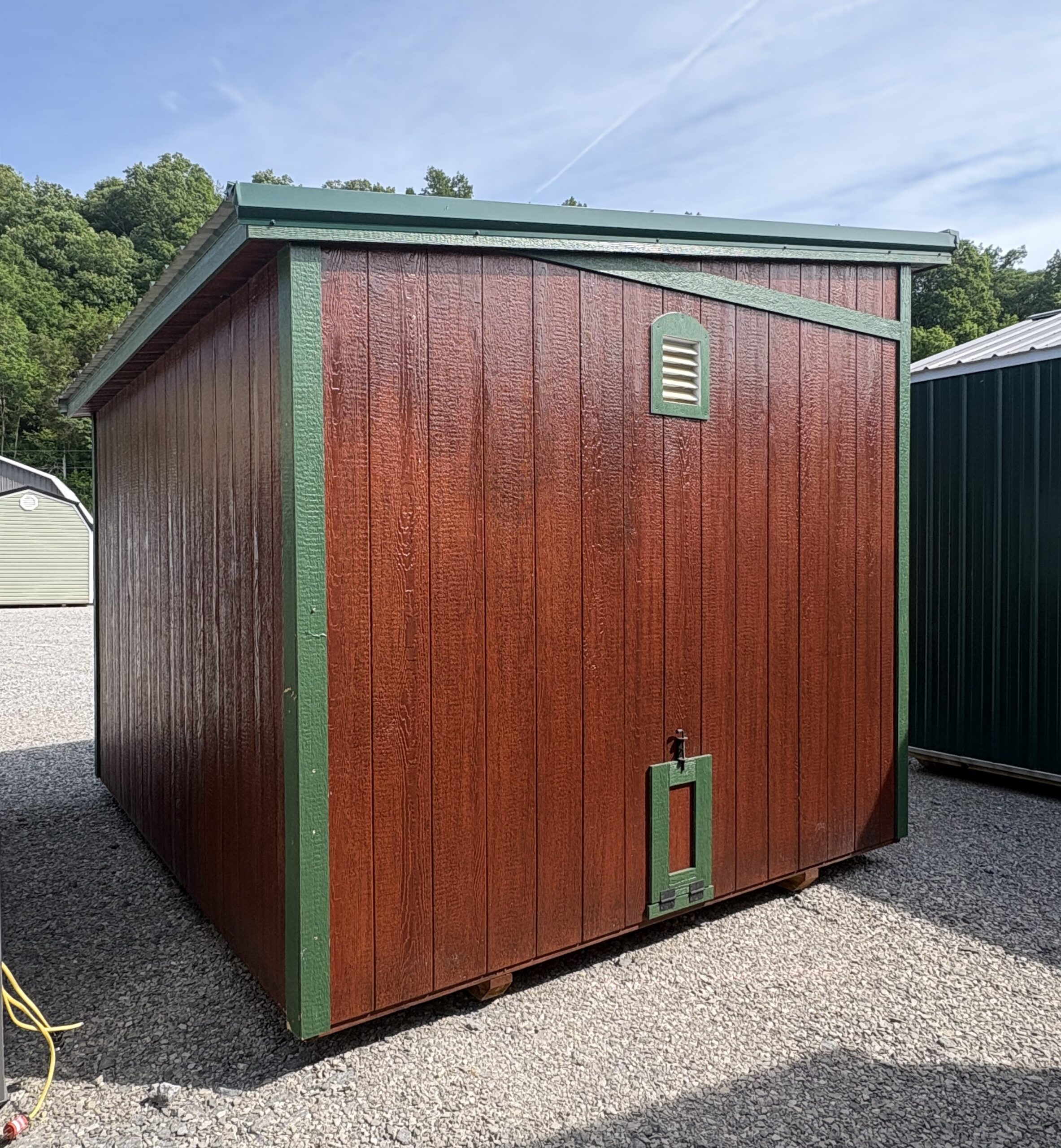 Red wooden storage shed with green trim and a small vent, set on a gravel lot with trees in the background.