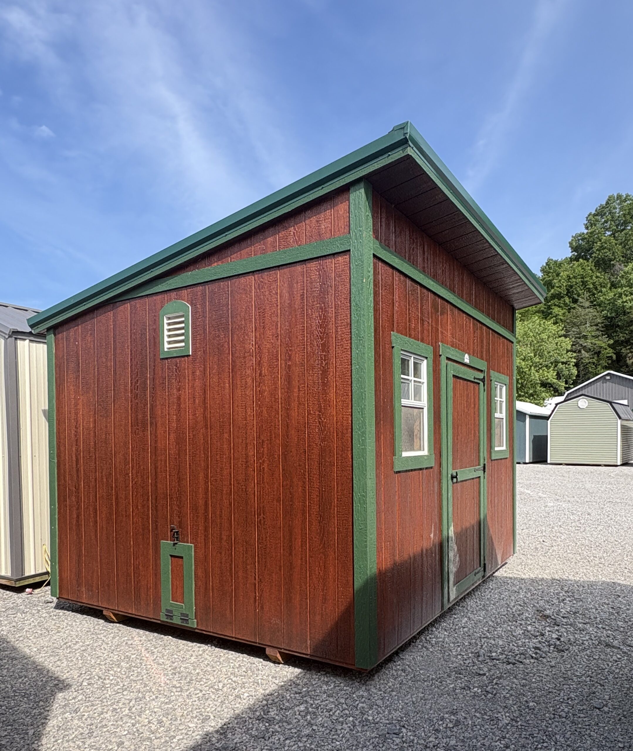 Corner view of a red wooden shed with green trim and white-framed windows on a gravel surface under a clear blue sky.