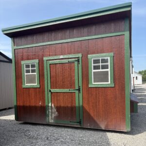 Small wooden shed with red walls and green trim, two windows and a door, on a gravel lot under a blue sky.