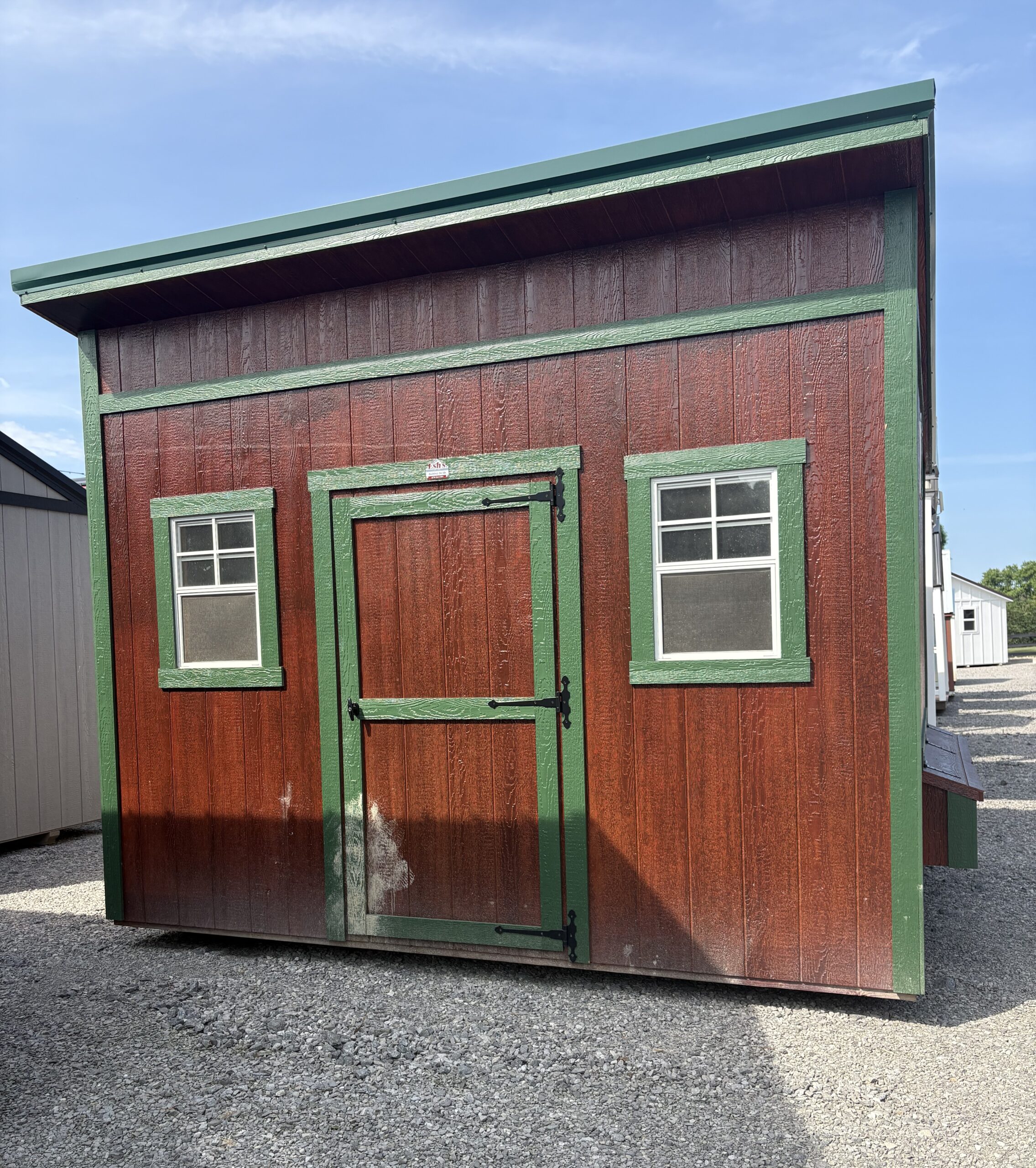 Small wooden shed with red walls and green trim, two windows and a door, on a gravel lot under a blue sky.