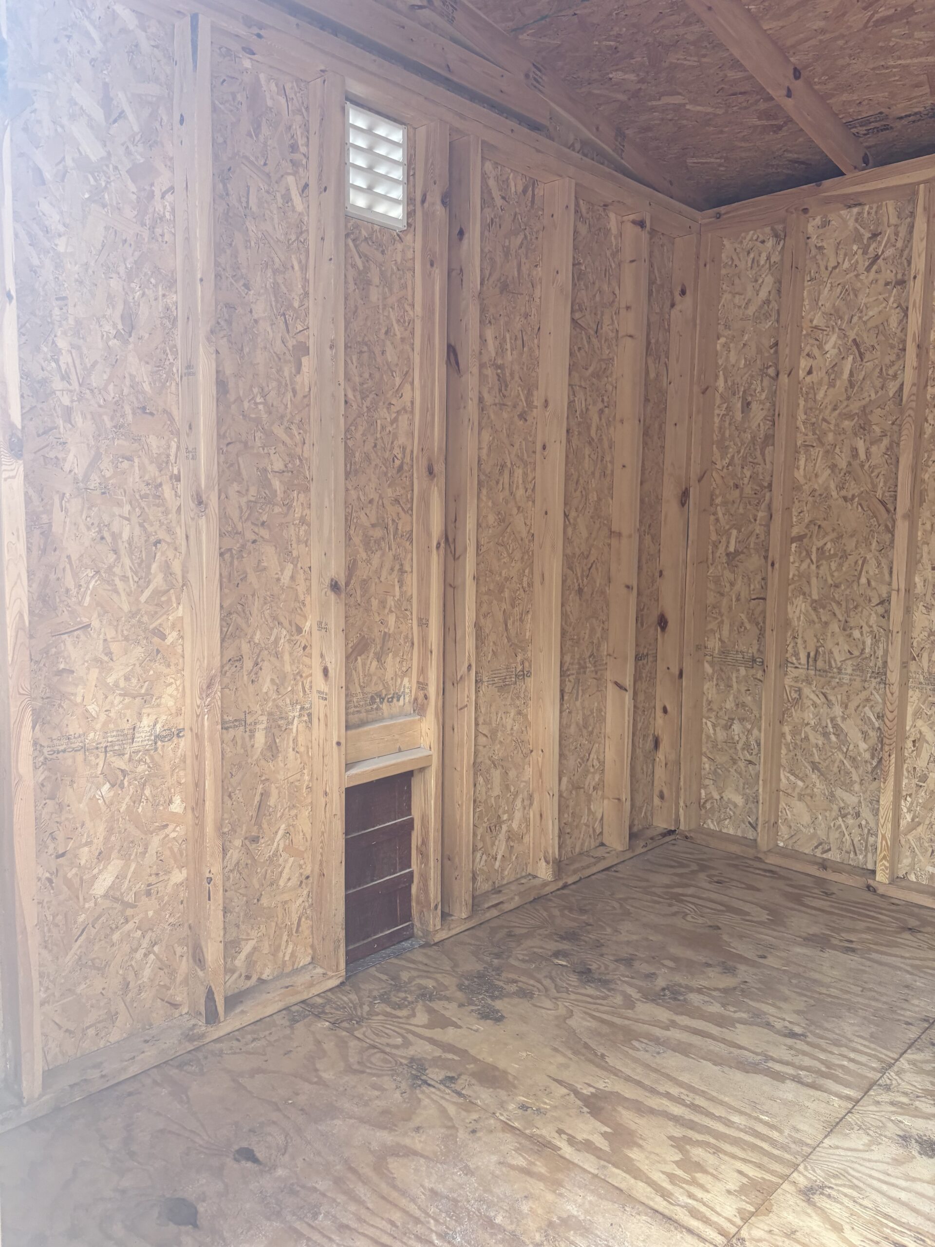 Unfinished wooden-framed interior with OSB walls and a small wall vent near the ceiling edge of a shed or utility room.