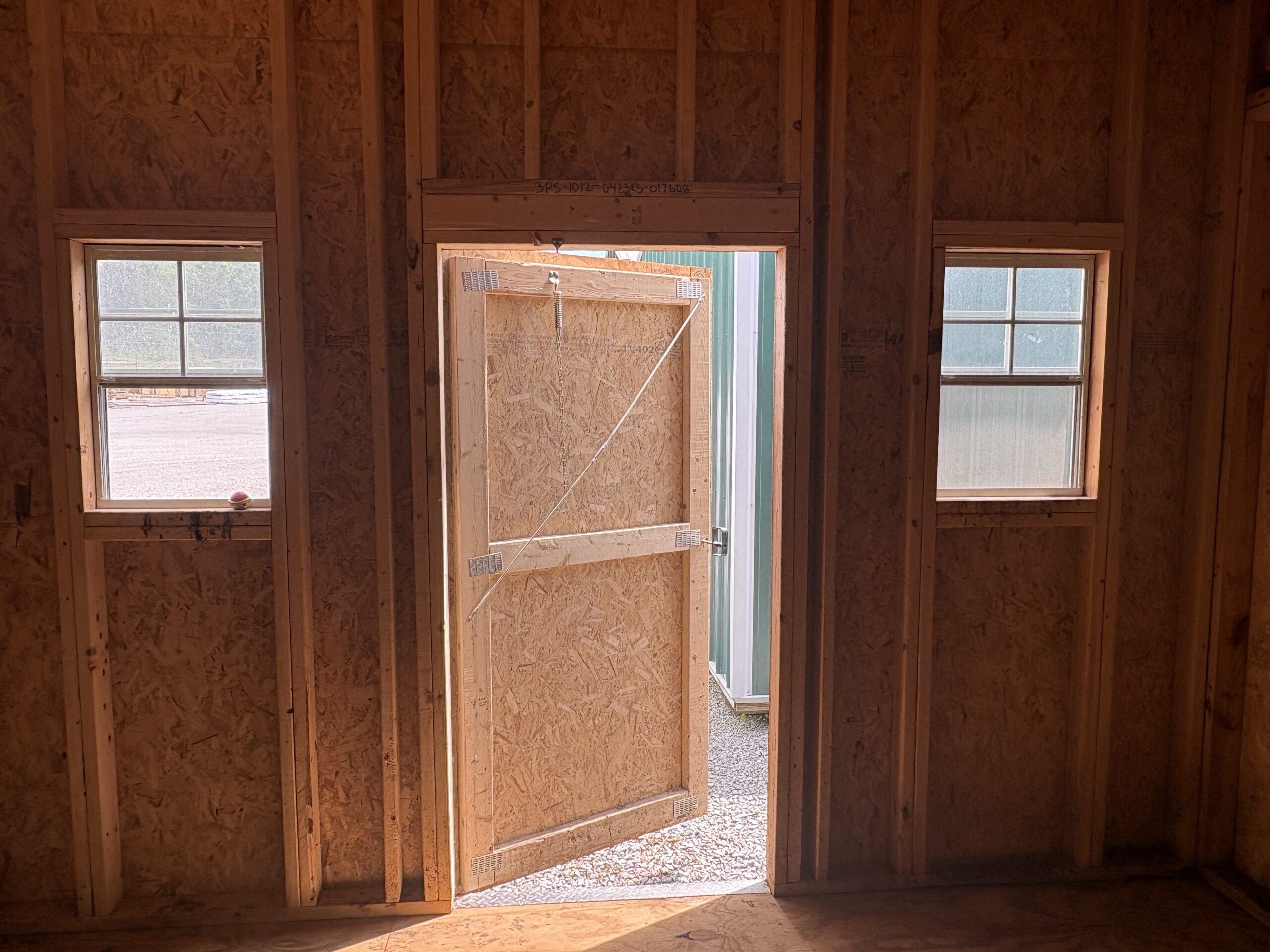 Interior view of a framed unfinished room with plywood walls, a doorway opening, and two windows to the left and right letting in daylight.