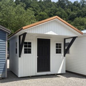White shed with a black front door, orange trim on the gabled roof, and dark diagonal supports on a gravel lot with trees behind.