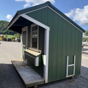 Small green wooden shed with white trim and a counter window on a gravel lot, with a playground in the background.