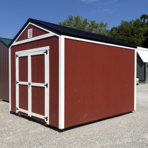 Red wooden shed with white trim and a black metal roof on a gravel lot, with a blue sky and trees in the background.