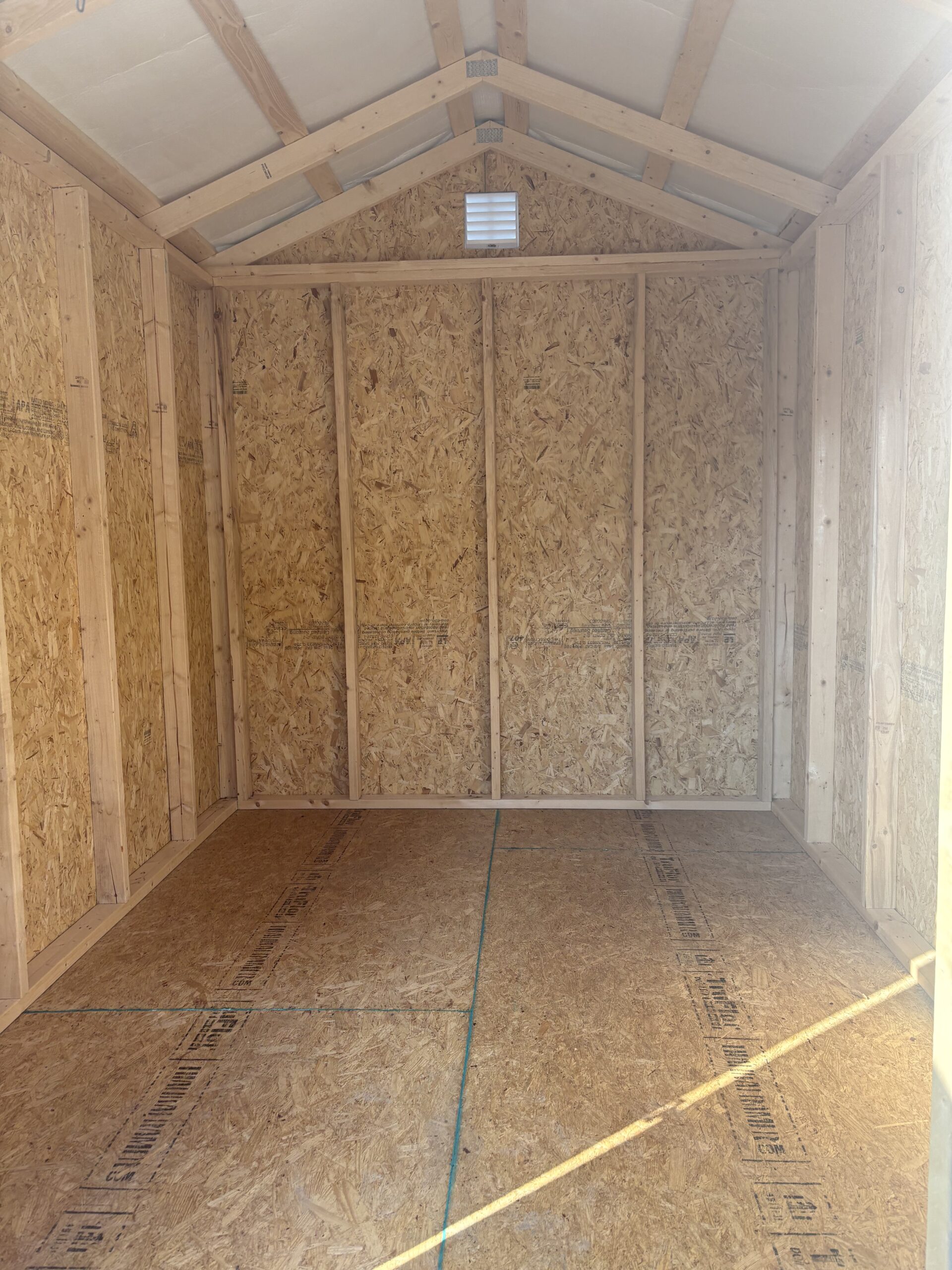 Interior of an unfinished wooden shed with exposed framing, OSB walls, and a pitched roof with a small vent near the peak while sunlight enters.