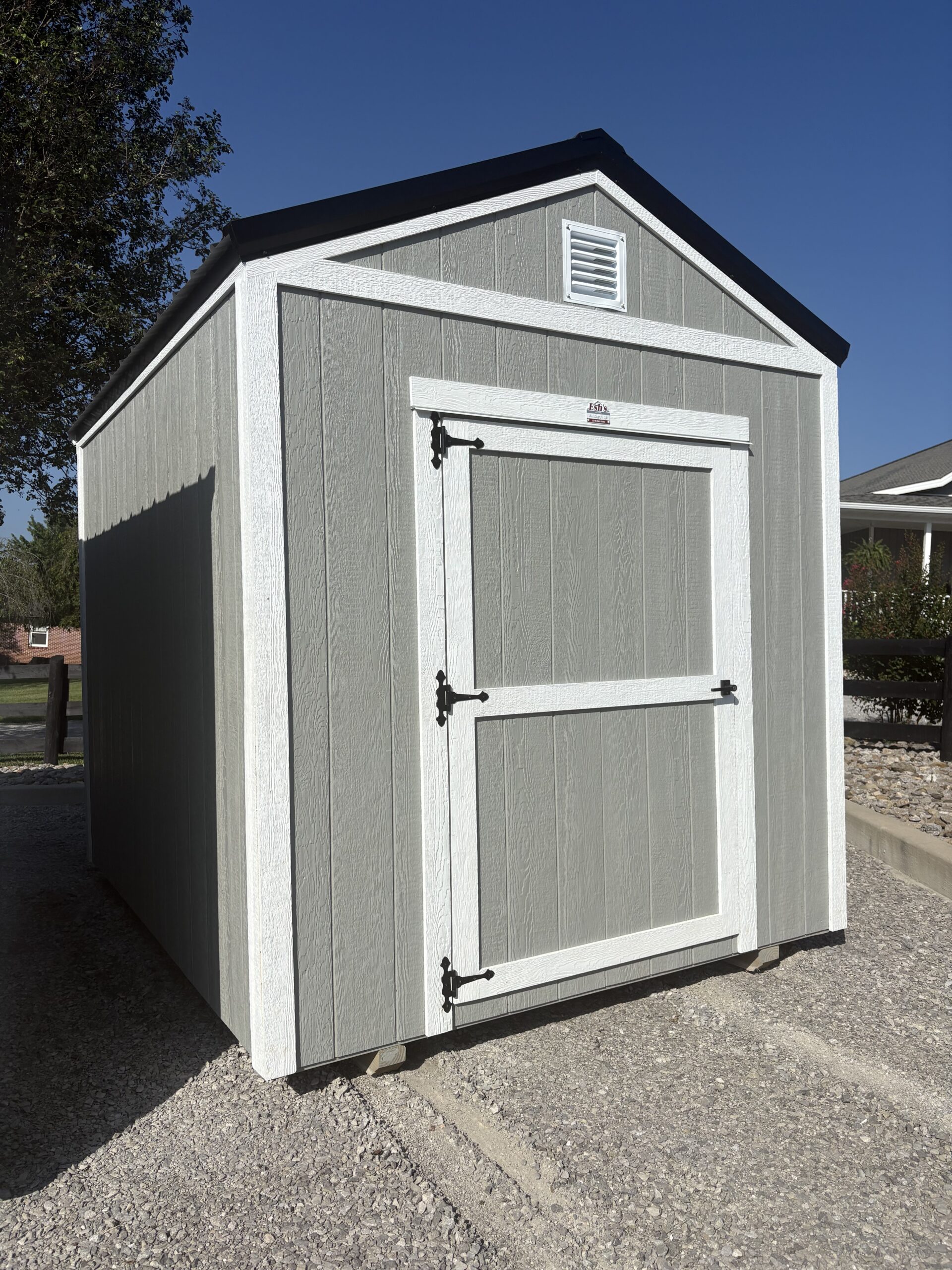 Small gray shed with white trim and a decorative double-door, located on a gravel surface under a blue sky. A roof vent is visible near the peak.