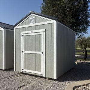 Gray storage shed with white trim and a small vent under the peaked roof, set on gravel with a tree in the background.