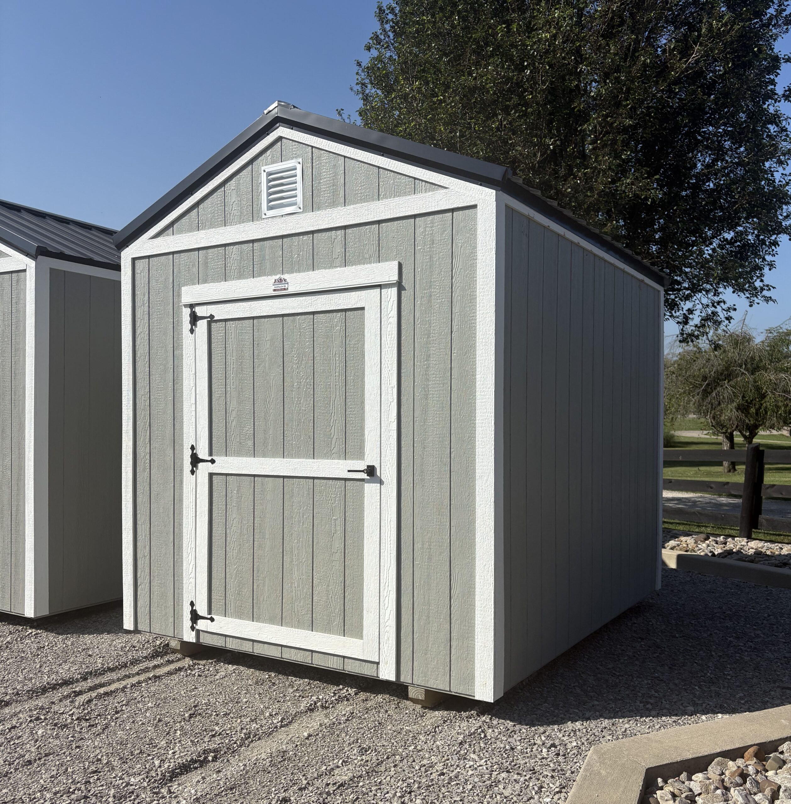 Gray storage shed with white trim and a small vent under the peaked roof, set on gravel with a tree in the background.