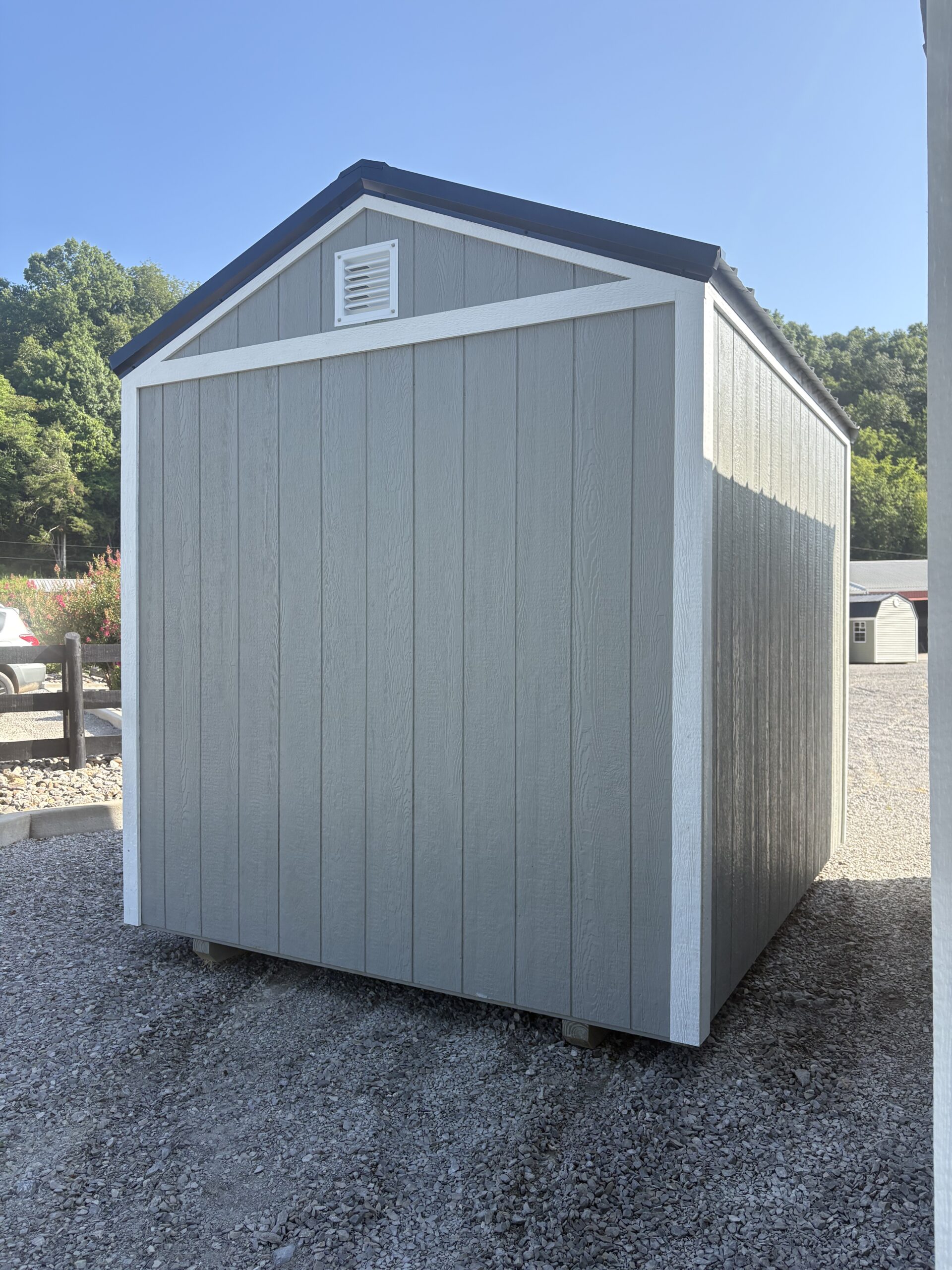 Gray storage shed with white trim and vent on the gable, set on gravel under clear blue sky.