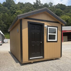 Small tan shed with black door and trim, white-framed window, outdoor light, on a gravel lot with trees in the background
