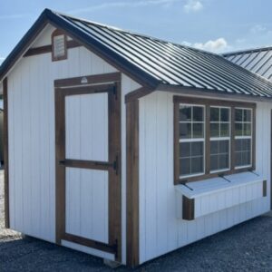 Small white shed with brown trim and a metal roof; door on the left and a large front window with a window box on the right.