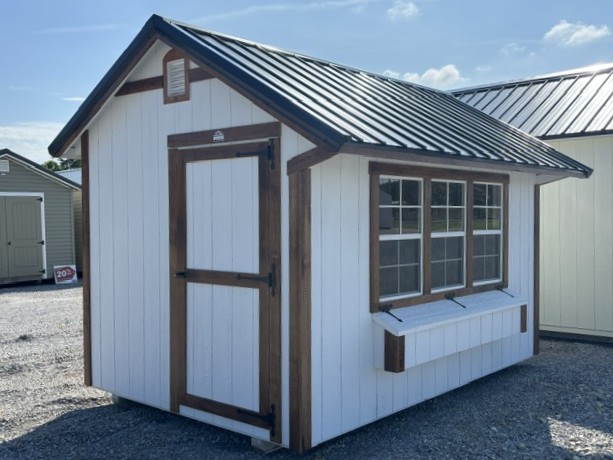 Small white shed with brown trim and a metal roof; door on the left and a large front window with a window box on the right.