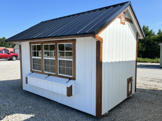Small white wooden shed with a dark metal roof, brown trim around the windows, and a window box, on a gravel lot.