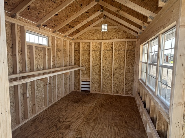Interior of a wooden shed under construction with exposed studs and rafters, a long workbench along the left wall, and windows along the right letting in daylight.