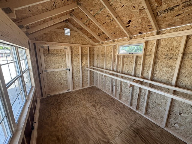 Interior of a partially finished wooden shed: exposed OSB walls and ceiling, plywood floor, a long workbench along the right wall, and windows with a door at the far end.
