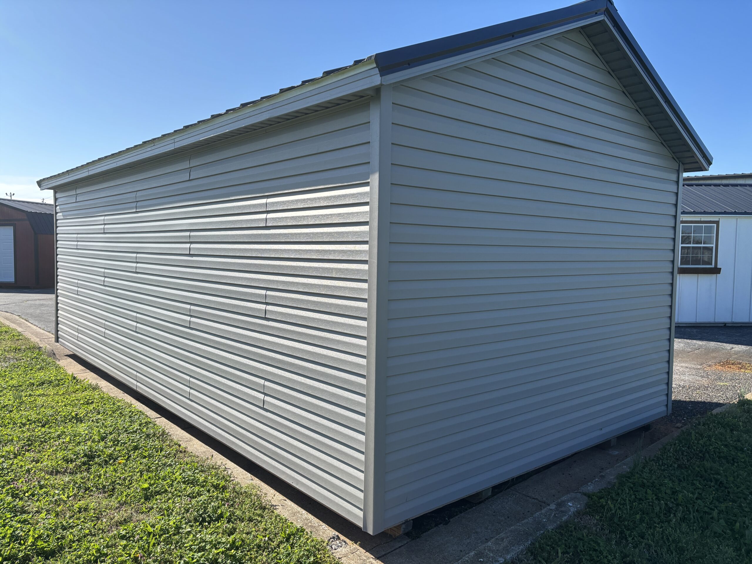 Metal storage shed with light gray horizontal siding and a pitched roof, set against a clear blue sky and surrounding buildings.