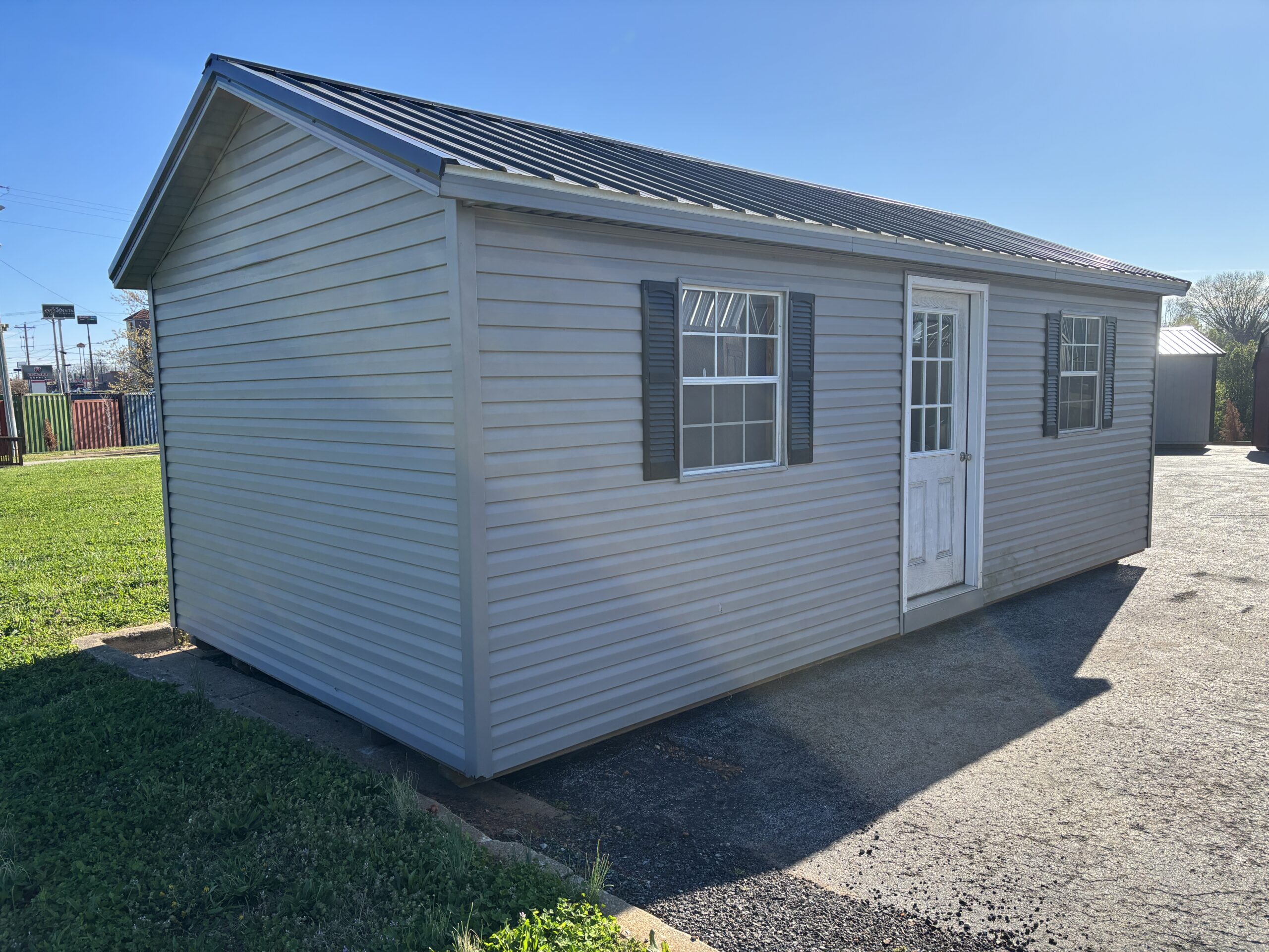 Small gray shed with a metal roof, two windows with shutters, and a white door on a paved area under a clear sky.