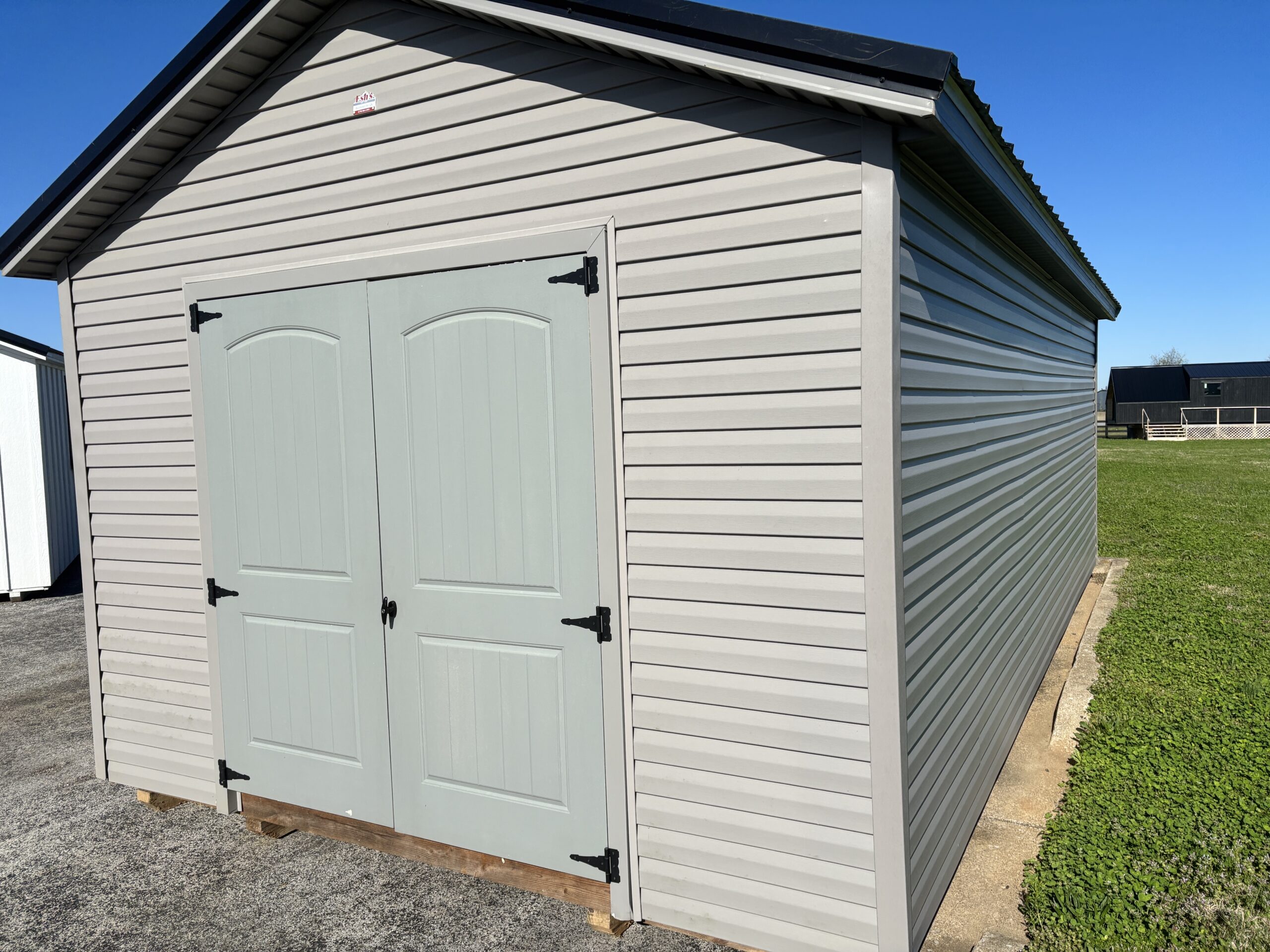 Gray shed with double doors and black hinges, set on a small wooden base on a concrete pad against a clear blue sky.