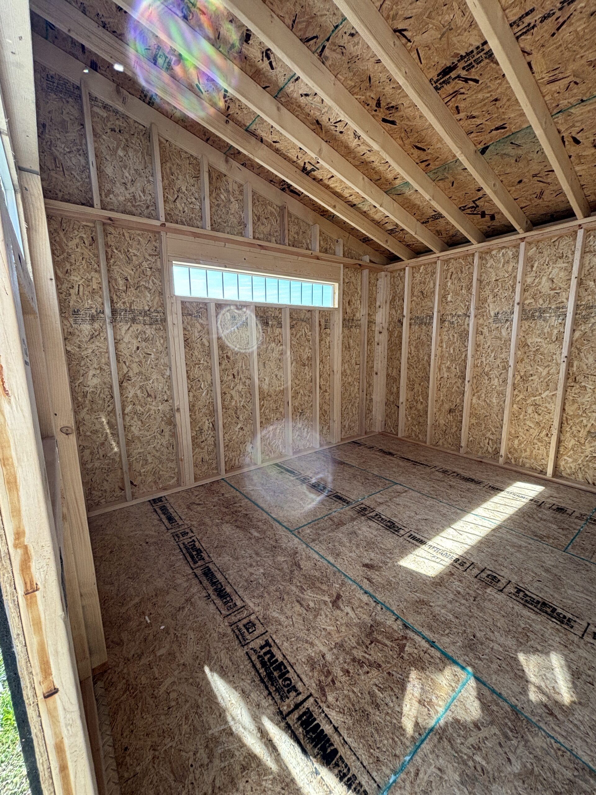 Interior of a new build with exposed wooden studs, OSB walls, and ceiling joists; sunlight through a narrow window.