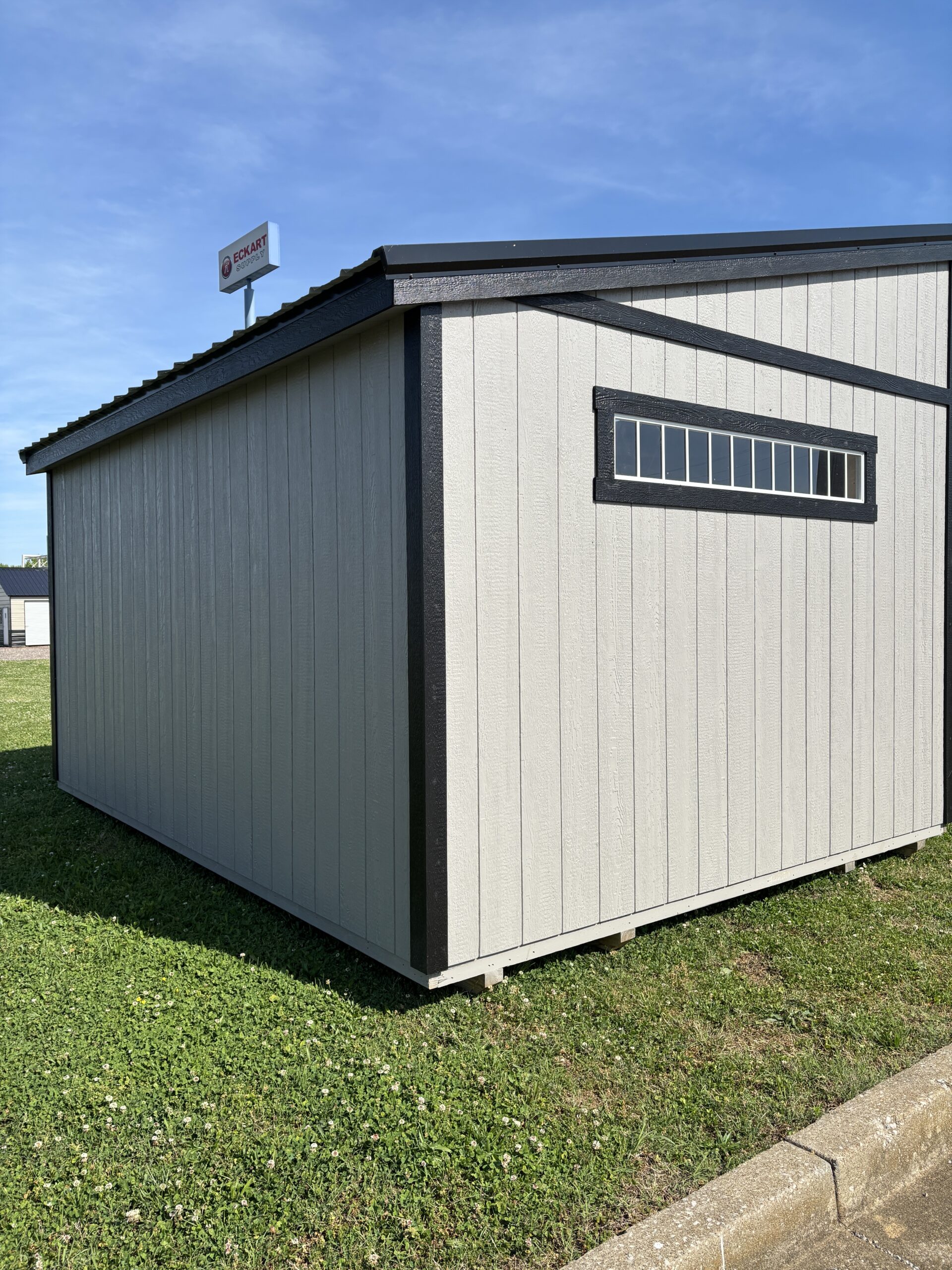 Gray storage shed with black trim and a small horizontal window, set on a grassy lot under a clear blue sky