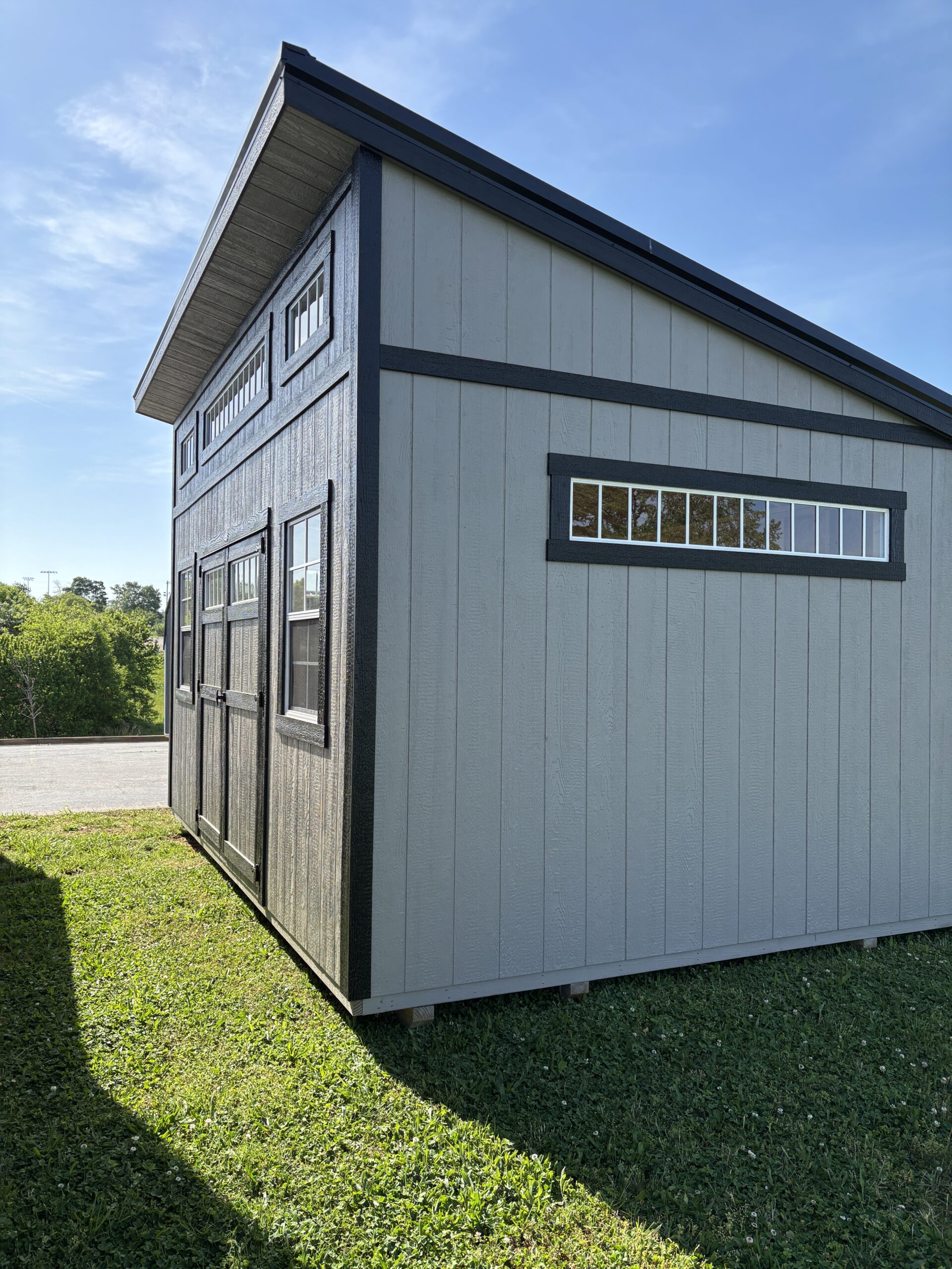 Gray wooden shed with black trim and a row of small upper windows, standing on a grassy lot under a blue sky, corner view.