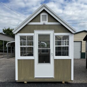 Front view of a small green shed with white trim, large glass door, and peaked roof; vented gable at the top. Educational: exterior on gravel with neighboring structures.