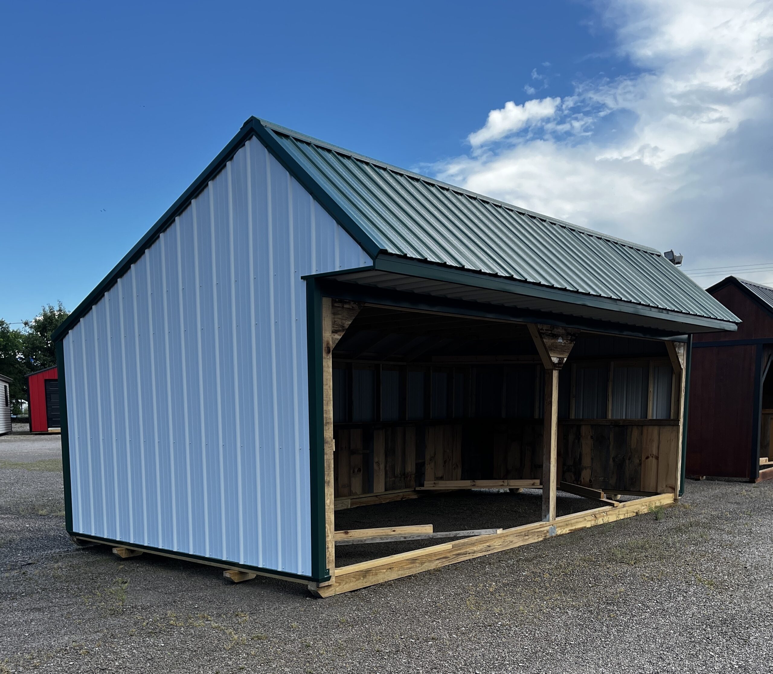 Blue metal shed with a corrugated roof and open front supported by wooden framing, under a blue sky in a gravel lot.
