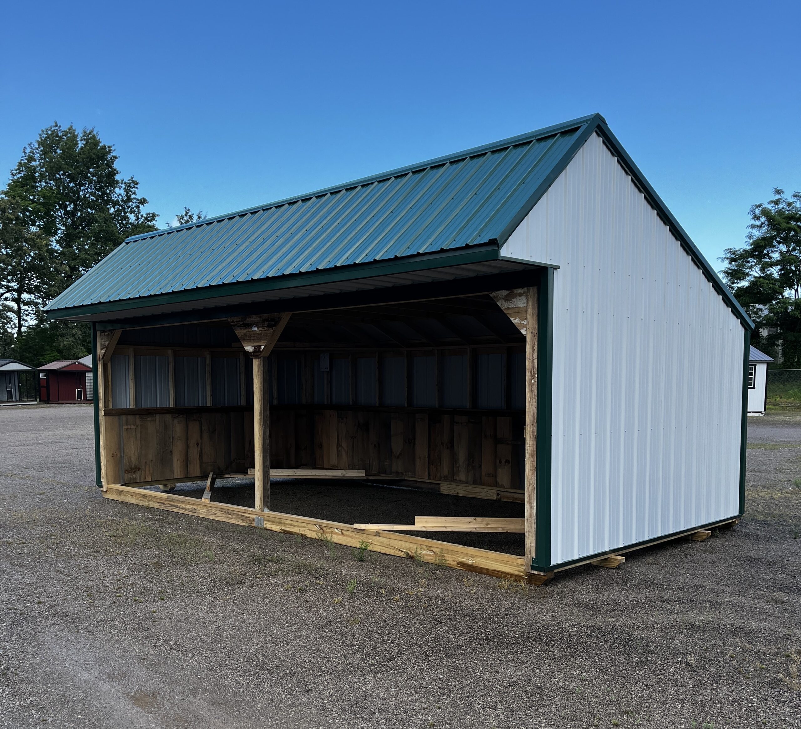 Open-sided wooden shelter with a blue-green corrugated metal roof in a gravel lot under a clear blue sky, with trees in the background.