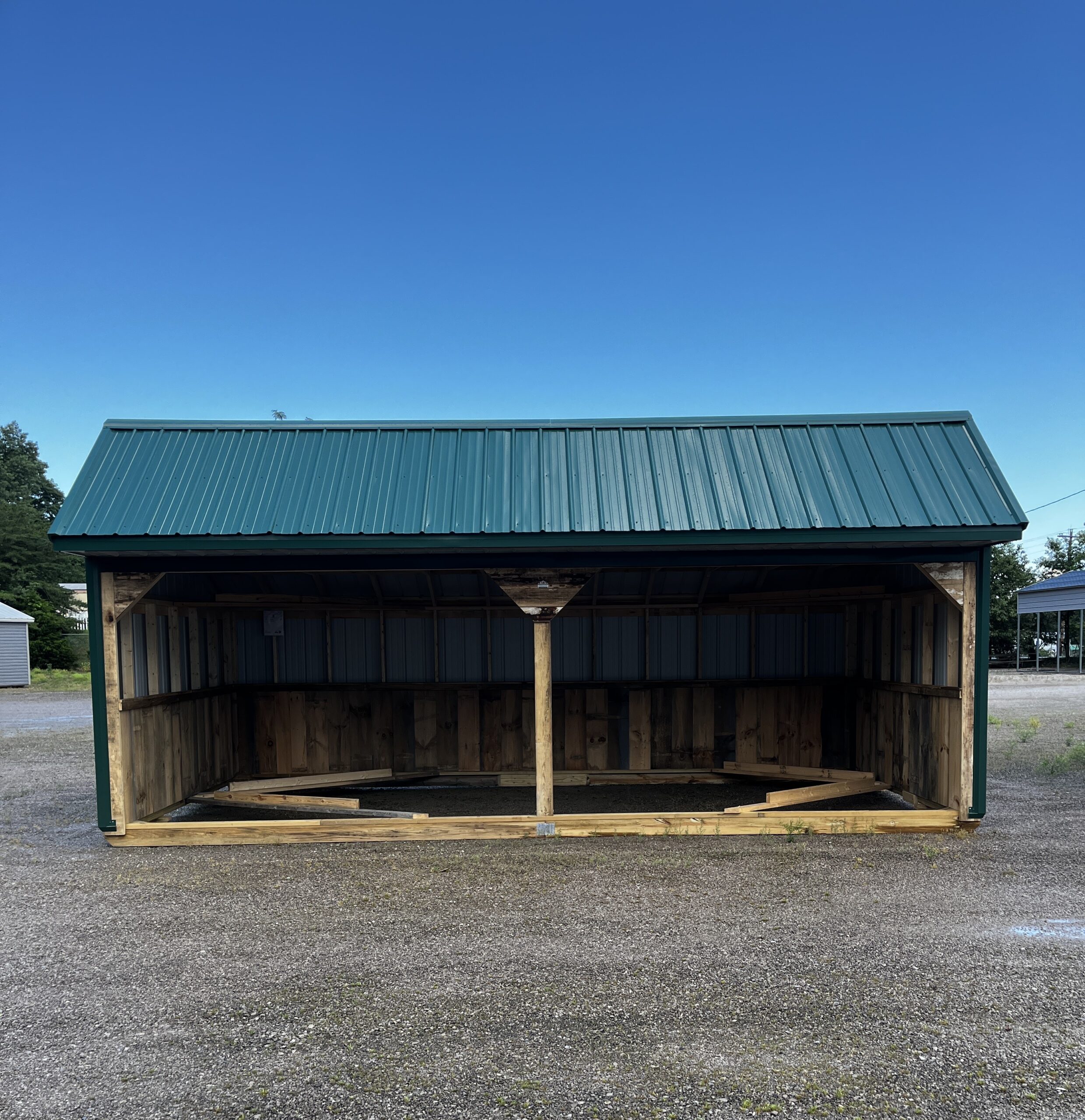 Open-sided carport or shelter with a teal metal roof over wooden framing, set on gravel under a clear blue sky