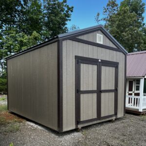 Gray shed with dark brown trim and vent on the gable, set on gravel with green trees and blue sky behind.
