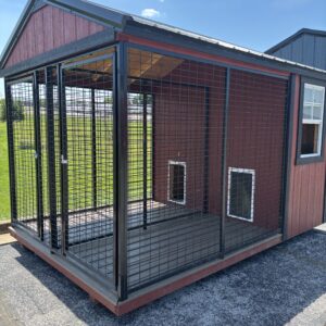 Outdoor metal dog kennel with wire mesh walls attached to a red wooden building on a concrete pad in bright sun