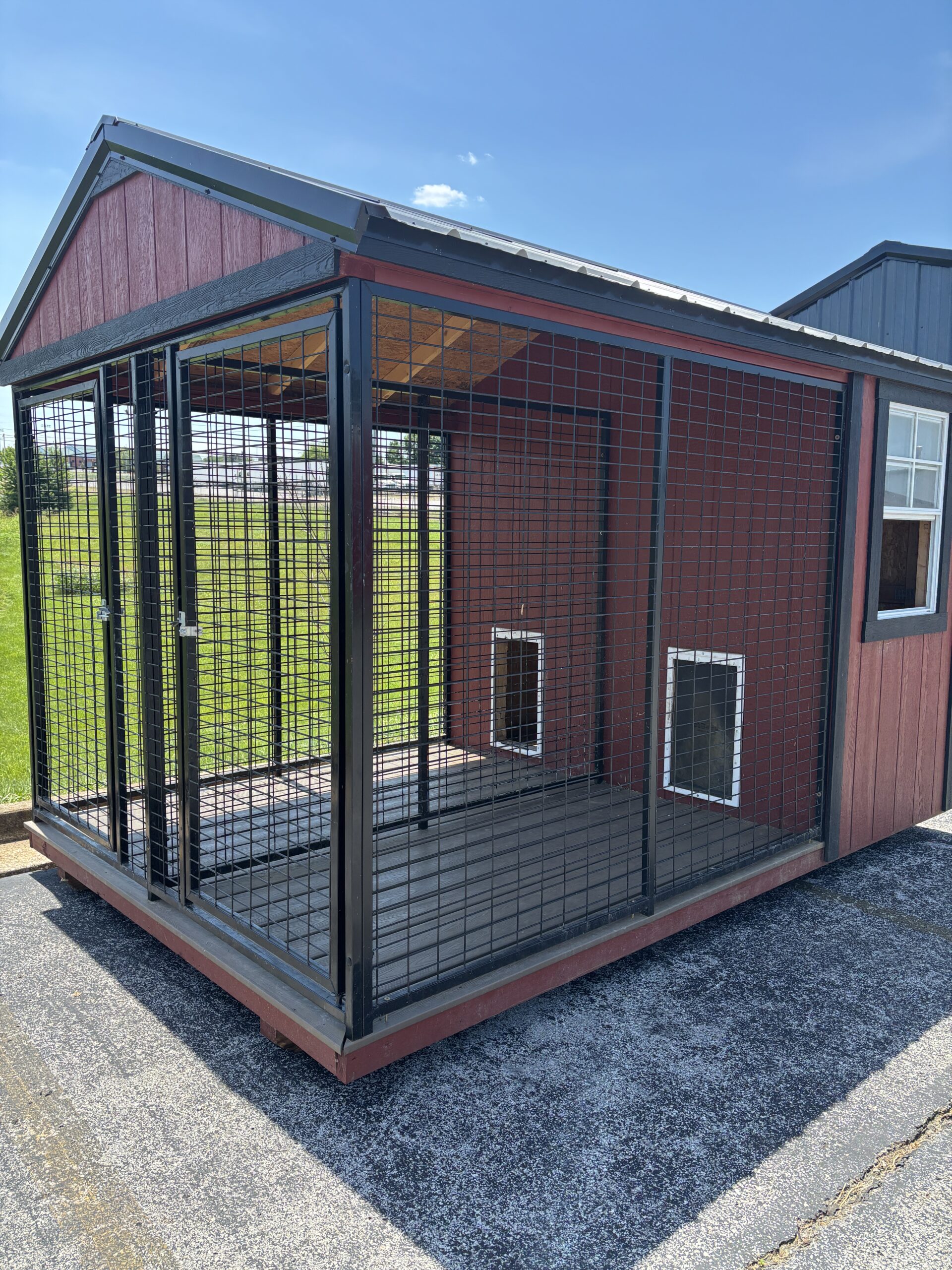 Outdoor metal dog kennel with wire mesh walls attached to a red wooden building on a concrete pad in bright sun