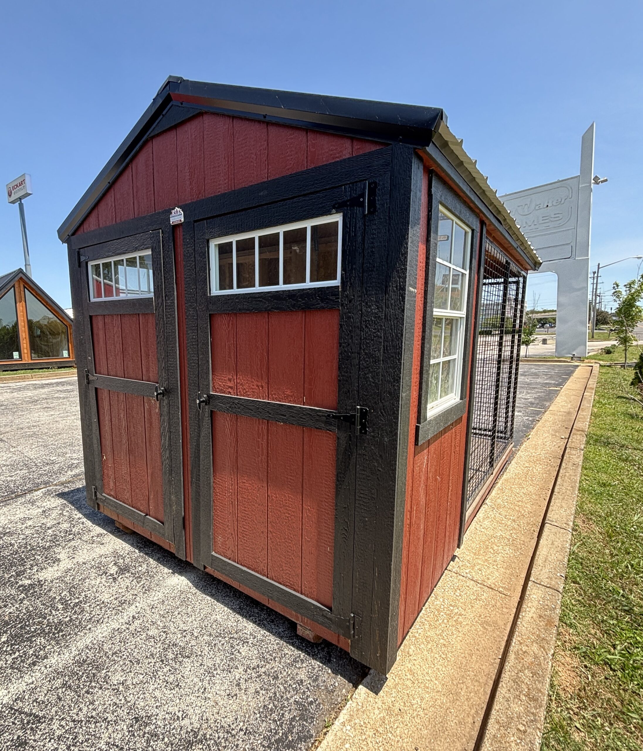 Red wooden kiosk or small shed with black trim and windows, outdoors on pavement with clear blue sky.