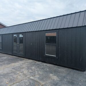 Black wooden shed with vertical siding, a tall standing-seam gray metal roof, and large glass doors/windows along the front, situated in a paved lot with a brick building nearby.